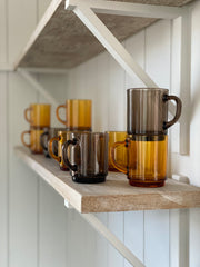 heavyweight french glass mugs with handles, some are amber, some are a cocoa brown. stacked on a shelf.