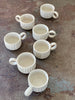 low mini mugs with a circular finger handle. the tiny cups are glazed white with a vertically carved stripe texture.