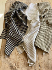 check print linen tea towels in black and white, natural and white and olive and white. the teatowels are displayed on an antique wooden table.